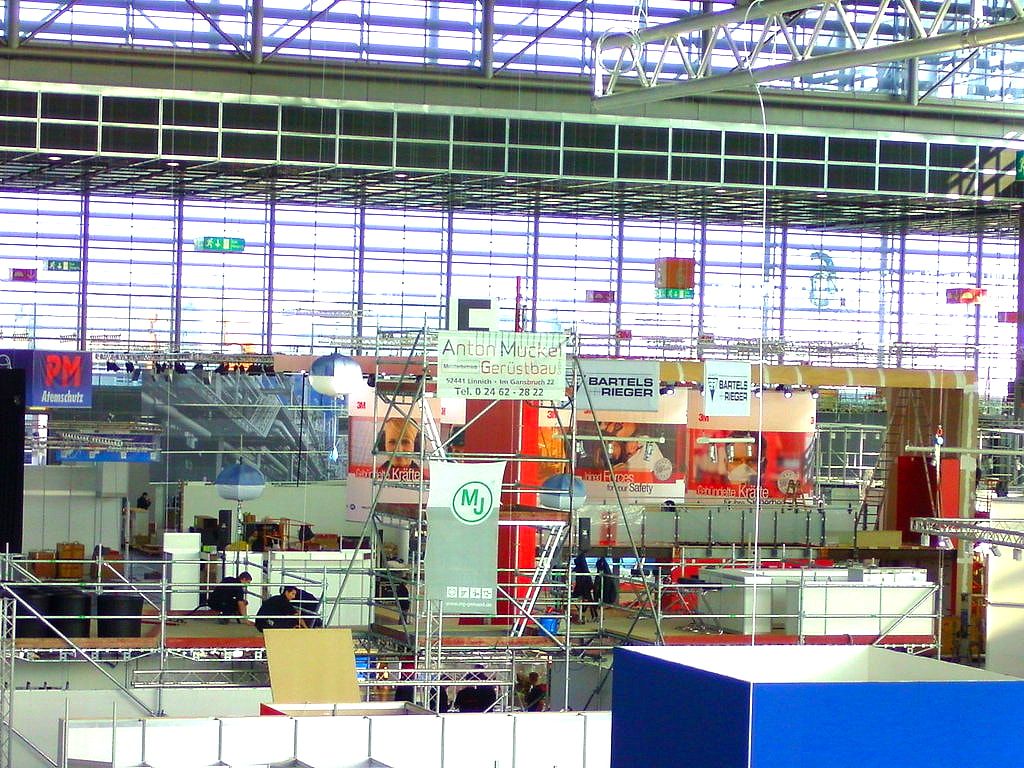 Weiter Blick durch eine Messehalle, Gerüstfelder und Standflächen unter der verglasten Dachkante